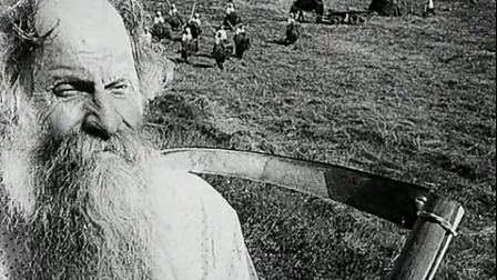 In the movie scene of Bezhin Meadow 1937, in a grassland with armed soldiers with sword and shields and horse at the back, in front is an old man who is serious, has bald top and white hair, beard and a mustache holding a thin scythe.