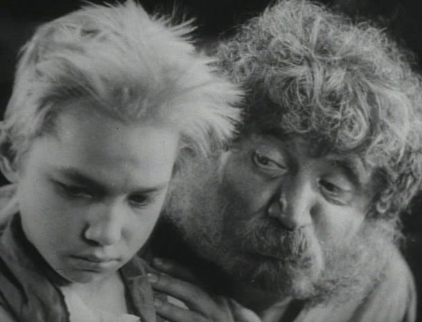 In the movie scene of Bezhin Meadow 1937, in black and white, from left Victor Kartashov is serious looking down, has white hair wearing a gray scarf and white top, at the right, Boris Zakhava is serious, looking at Victor’s face, with his left hand on the shoulder has white curly white hair a beard and a mustache wearing a gray shirt.