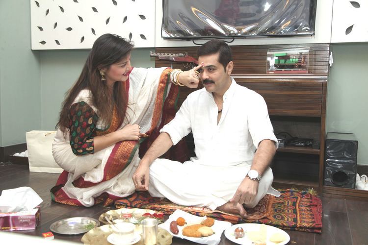 In a room with a flat screen tv, cabinets, from left, a woman is smiling, crouching while applying tikka on the man's forehead, has long black hair wearing earring, bracelet, and white saree, at the right a man is serious, sitting, has black hair mustache, wearing a silver watch a white top and white pants, in front is a plates with foods.