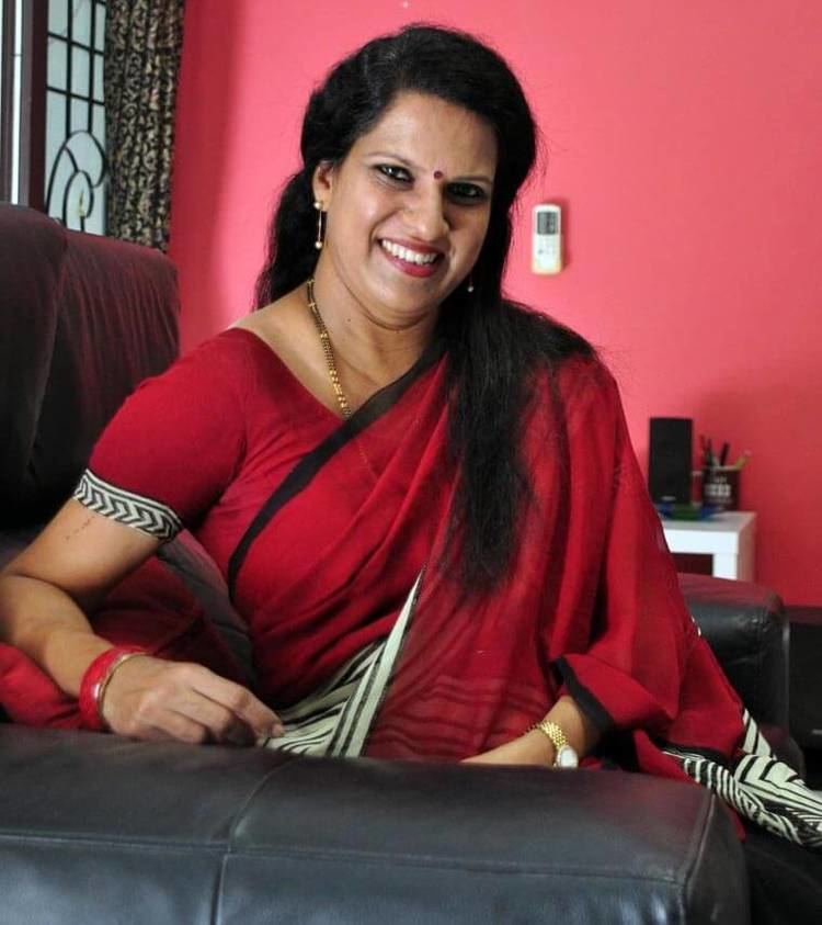 Bharathi Baskar smiling while sitting on a black couch and wearing a red and black dress, red bracelet, wristwatch, necklace, and earrings