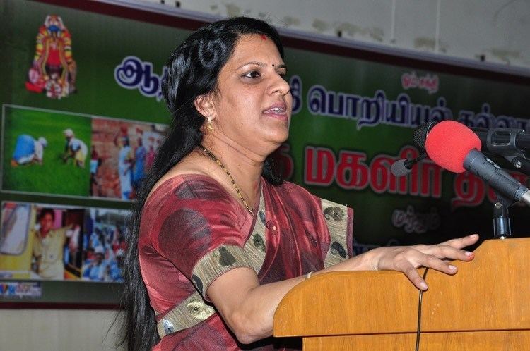 Bharathi Baskar giving a speech while wearing a brown and maroon dress, necklace, and earrings