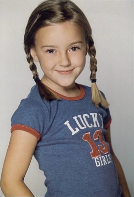 Young Billi Bruno smiling with braided blonde hair and wearing a blue shirt with red and white print.