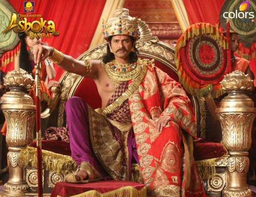 Bindusara Maurya is seriously sitting down on the Throne, has black hair, wearing a turban made of gold and precious gems such as rubies, diamonds, emeralds, and sapphire jewelry, red choghah, a violet dhoti pants, and gold jutti.