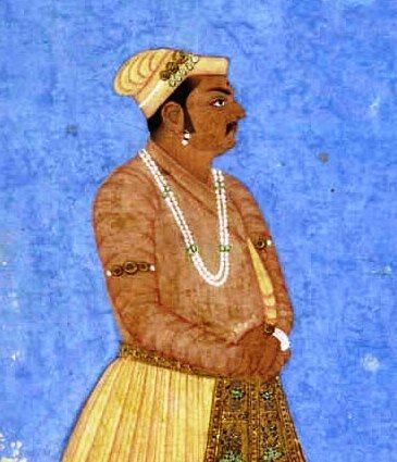 A painting of Birbal looking serious facing to the side with a black tika on the forehead, wearing headgear with jewels in it, a bead necklace, bands on the arms, earrings, a bracelet, and orange pants under a yellow skirt