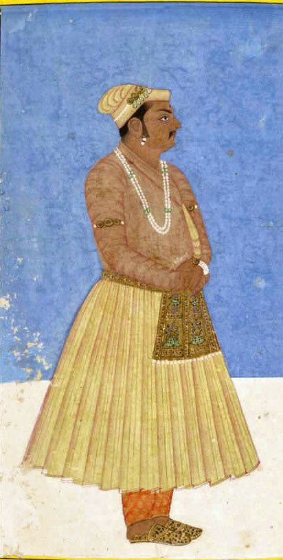 A painting of Birbal looking serious facing to the side with a black tika on the forehead, wearing headgear with jewels in it, a bead necklace, bands on the arms, earrings, a bracelet, traditional shoes long sleeve and orange pants under a yellow skirt