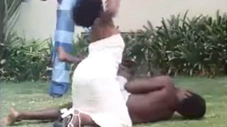 A black man and woman, a scene from the 1974 Blaxploitation film written and directed by Jamaa Fanaka, Black Sister's Revenge. The woman is punching the man on the ground with her arms in the air, and plants in the background with white sandals in a white tube dress. The man is topless, wearing a white bottom.