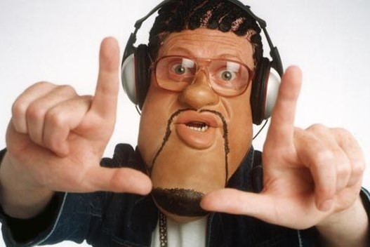 Leigh Francis impersonates Craig David on the set of Bo' Selecta! while doing a loser hand sign, with curly hair, wearing headphones, eyeglasses, and a denim polo shirt over a white shirt.
