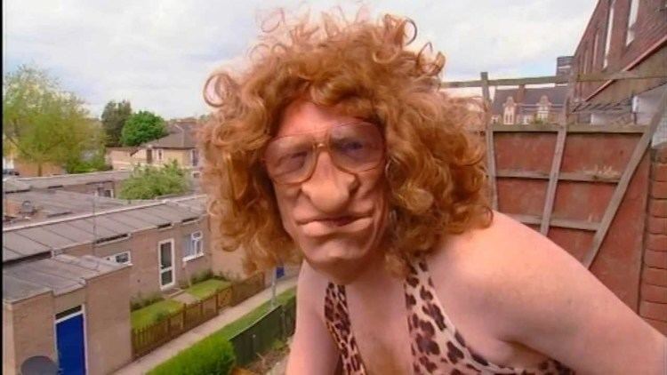 Leigh Francis impersonates Mel B. on the set of Bo' Selecta!, with a grumpy face, wearing a curly blonde wig, eyeglasses and animal printed top.