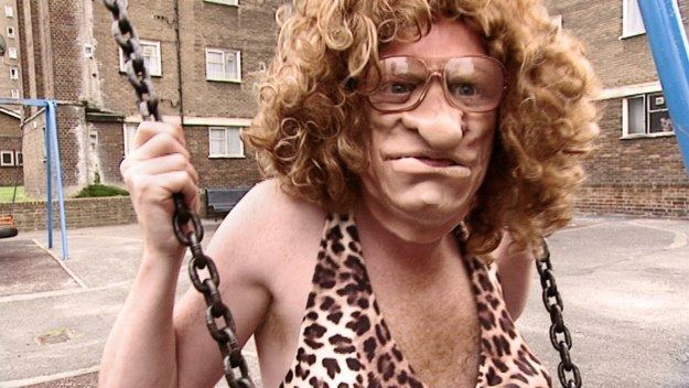 Leigh Francis impersonates Mel B. on the set of Bo' Selecta! while riding on a swing, with a grumpy face, wearing a curly blonde wig, eyeglasses and animal printed top.