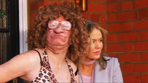 Leigh Francis impersonates Mel B., with a grumpy face while Patsy Kensit is smiling. Leigh wearing a curly wig and animal printed top while Patsy wearing a purple coat in a scene from Bo' Selecta!, a British television sketch show.