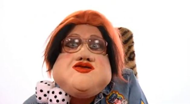 Leigh Francis impersonates Trisha Goddard on the set of Bo' Selecta!, with a dice near his neck, with a swollen face, red hair, wearing eyeglasses, and a denim shirt on the set of Bo' Selecta!, a British television sketch show.