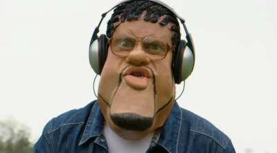 Leigh Francis impersonates Craig David on the set of Bo' Selecta!, with curly hair, wearing headphones, eyeglasses, a denim polo shirt over a white shirt.