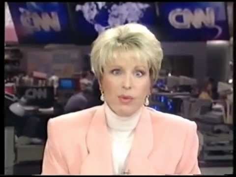 In CNN scene, at the back is a studio office with staffs and electronics, screen on broadcasting, CNN and a world map with blue background, in front, Bobbie Battista is serious, eyebrow up, mouth half open, has blond hair wearing a pearl teardrop earring, white turtleneck shirt under a pink coat.