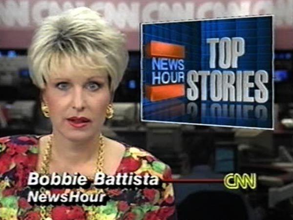 In a News scene of CNN, at the back is a studio office and electronics screen on broadcasting, CNN with red background, in front, Bobbie Battista is serious, mouth half open, has blond hair wearing a gold earring, gold necklace and red floral printed blouse with a word with her name “Bobboe Battista” “NewsHour” at the top right is a box with introduction of the NewsHour Top Stories written on it, at the right bottom is the CNN logo.