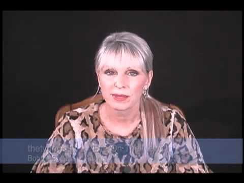 In a dark gray background, Bobbie Battista is serious, sitting in a brown wooden chair, has white hair wearing silver earrings and an animal printed blouse.