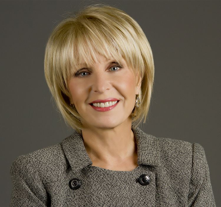 In a gray background Bobbie Battista is smiling, has blond short hair, gray eyes, wearing red lipstick, gold earrings, and a gray winter coat with large black buttons.