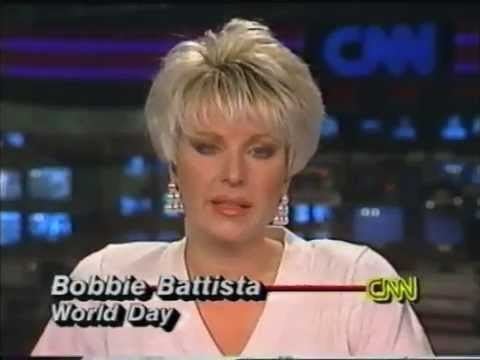 In a News scene of CNN, at the back is a studio office and electronics screen on broadcasting, CNN with blue background, in front, Bobbie Battista is serious, mouth half open, has blond hair wearing a pearl chandelier earring, and  a pink dress with a word with her name “Bobboe Battista” “world day” at the right is the CNN logo.