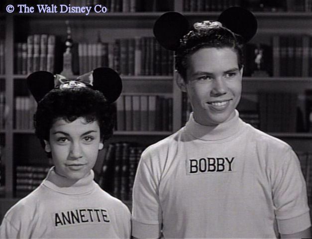 Annette Funicello and Bobby Burgess are smiling. Both are wearing a mickey mouse headband and a white shirt with their names.