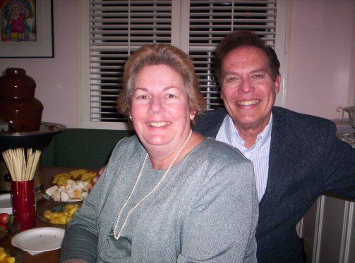 Bobby Burgess and Kristie Floren Burgess are smiling while sitting on a couch. Bobby wearing a gray coat over white striped long sleeves while Kristie wearing a necklace and a gray long sleeves shirt.
