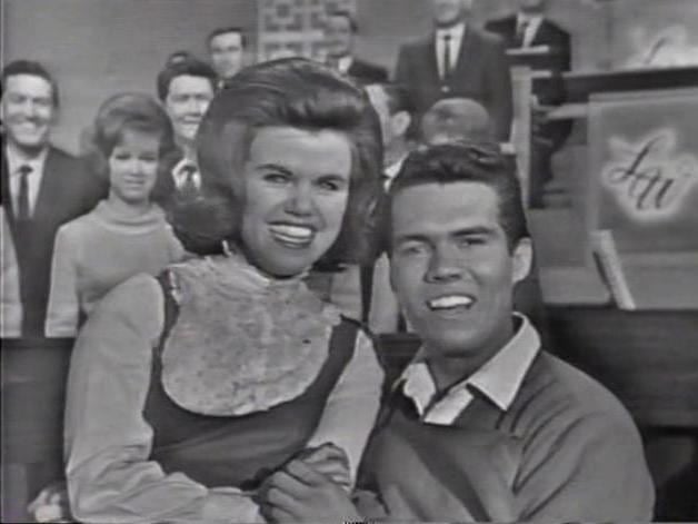 Cissy King and Bobby Burgess are smiling while holding hands. Cissy with short hair and wearing a jumper dress over a white long sleeves shirt while Bobby wearing a polo shirt with a white collar.