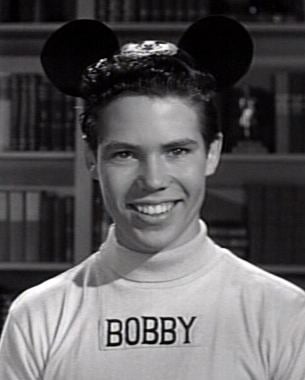 Bobby Burgess smiling, wearing a mickey mouse headband and a white shirt with his name.