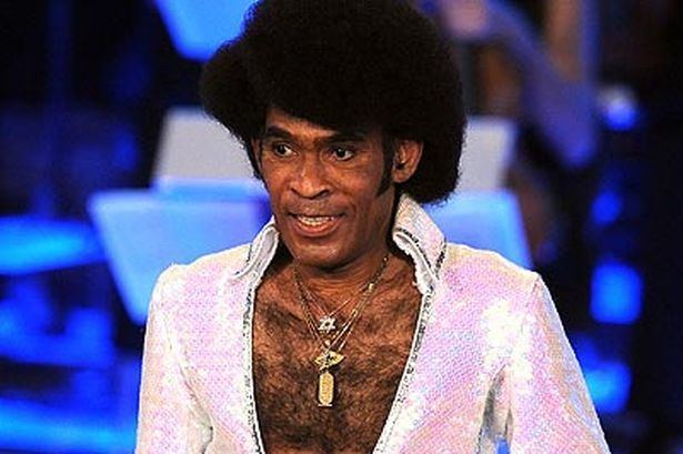 Bobby Farrell smiling on stage during a concert wearing a white bejeweled collared shirt and some necklaces.