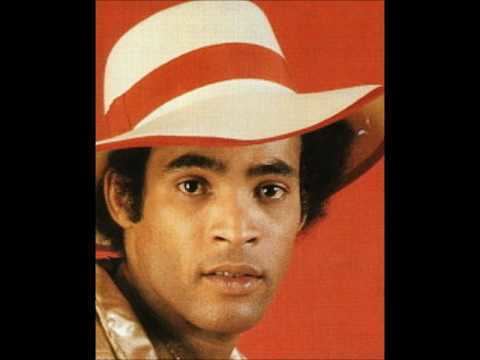 Bobby Farrell smiling during a music video and wearing a white hat and a pink jacket.