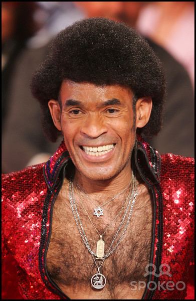 Bobby Farrell smiling on stage during a concert wearing a red bejeweled collared shirt and some necklaces.