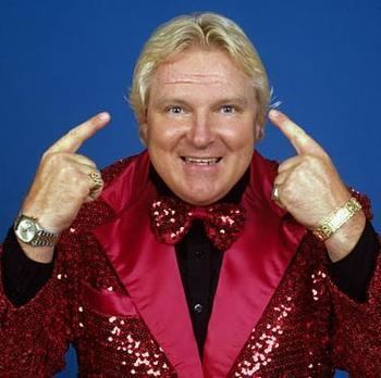 Bobby Heenan smiling while pointing his finger to his head and wearing a red coat with sequins, black long sleeves, bow tie, wristwatch, and bracelet