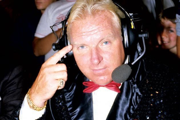 Bobby Heenan pointing his finger to his head while wearing a headset, black coat, white long sleeves, red bow tie, gold bracelet, and ring