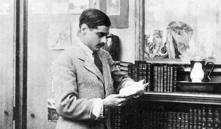 Arthur Edward "Boy" Capel reading a book while leaning on the bookshelves, with a mustache, and wearing a coat and tie.