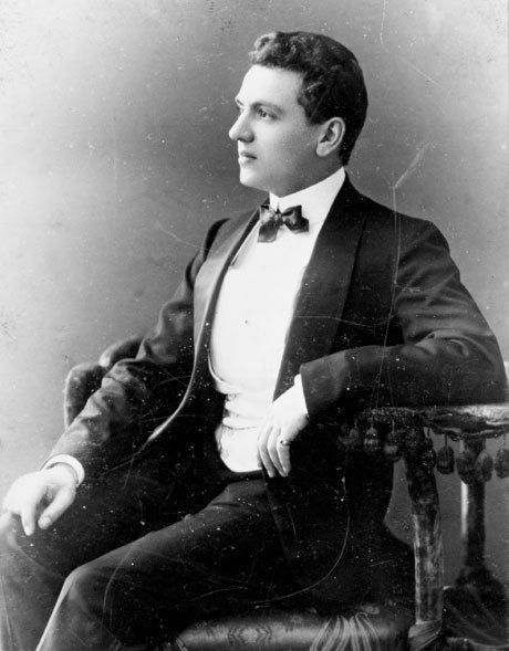 Arthur Edward "Boy" Capel with a serious face while sitting on a chair, wearing a black coat over white long sleeves and a black bowtie, and black pants.