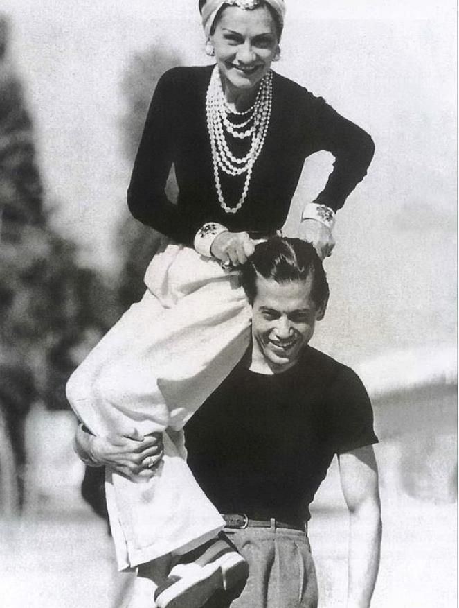 Arthur Edward "Boy" Capel smiling while carrying Coco Chanel. Boy wearing a black shirt while Coco wearing earrings, a pearl necklace, a black long sleeve top, and white pants.