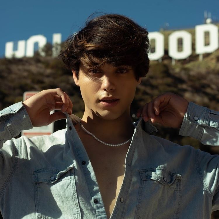 Brandon Cardoso with a serious face while holding the collar of his denim shirt, and wearing a pearl necklace.