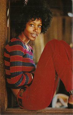 Brenda Sykes sitting down and smiling while wearing a black and red striped sweatshirt and red pants.
