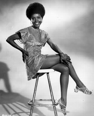 Brenda Sykes smiling and sitting down with her hand in her waist while wearing a glittering dress.