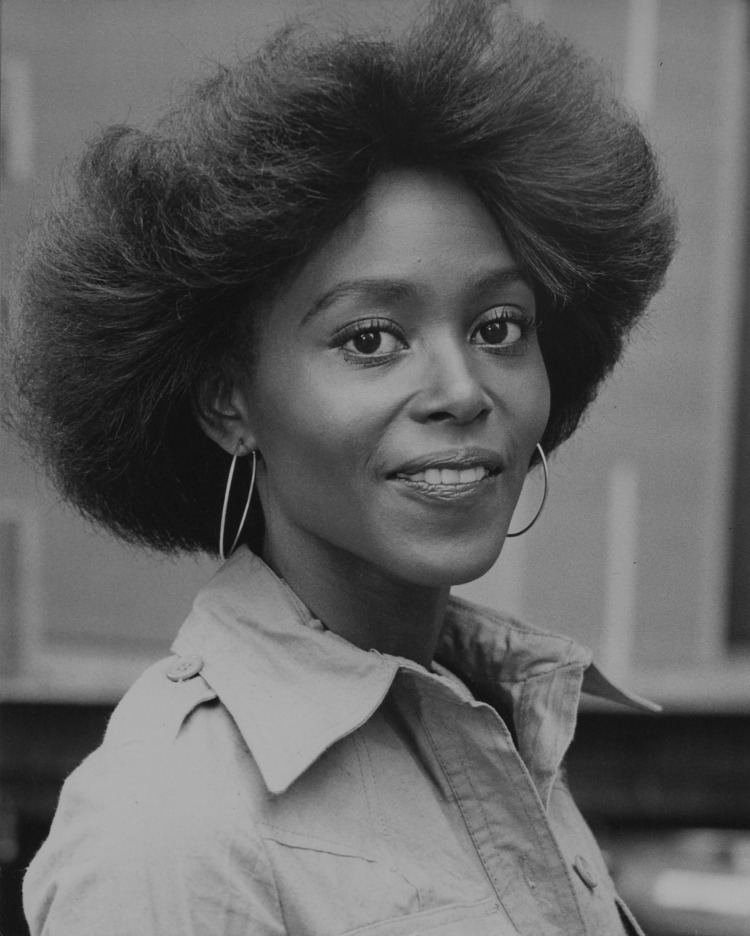 Brenda Sykes smiling with thick hair and wearing a white dress and large earrings.