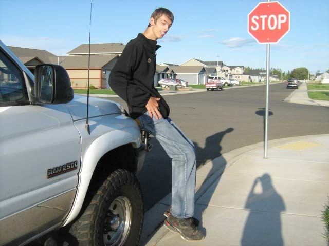 Brenden Adams smiling while sitting on a car, wearing a black jacket, blue jeans, and black shoes.