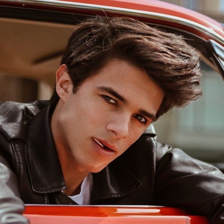 Brent Rivera biting a toothpick while leaning on his car's window and wearing a white shirt under a black leather jacket