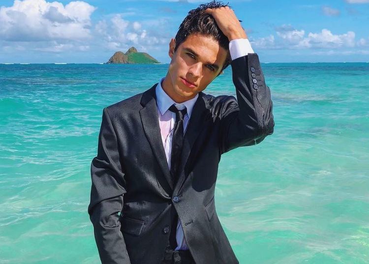 Brent Rivera looking fierce in a standing pose at the sea and holding his hair while wearing a white sleeve under a black suit and a black necktie