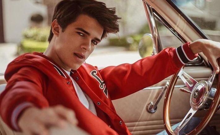Brent Rivera biting a toothpick with a serious look and holding his car's steering wheel while wearing a shirt under a red jacket