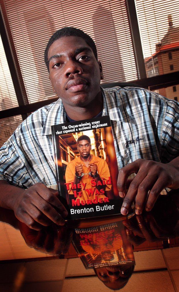 Brenton Butler holding the book "They Said It Was Murder" with a serious face while wearing a white, blue, black checkered polo