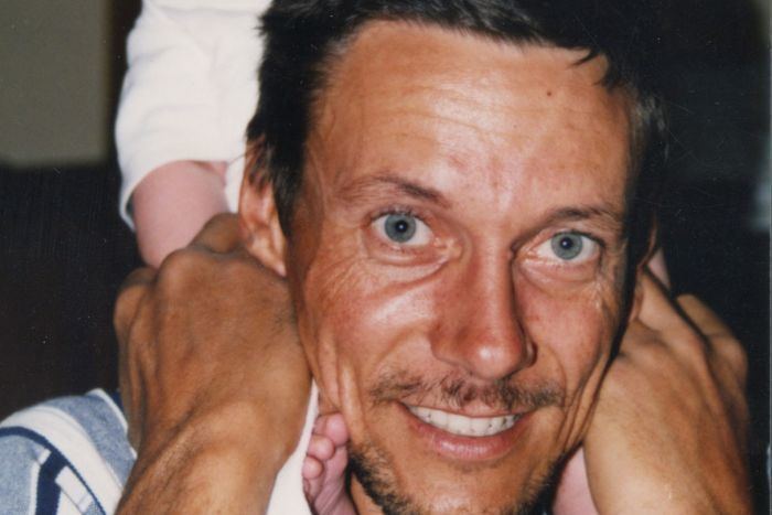 Brett Peter Cowan is smiling, holding a baby wearing white clothes on the back of his head, has black hair, gray eyes wearing a striped blue shirt.