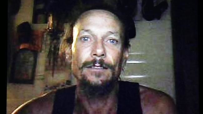 In a room with picture frames and wall decorations on the white wall. Brett Peter Cowan is serious, has black hair, mustache and beard, wearing a black tank top.