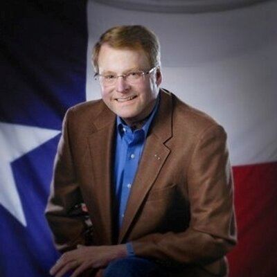 Brian Birdwell smiling while his arm on his leg, with a Texas flag on the background, wearing eyeglasses, a brown coat over blue long sleeves.