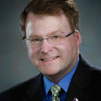 Brian Birdwell smiling, wearing eyeglasses, a black coat over blue long sleeves, and a green checkered tie.