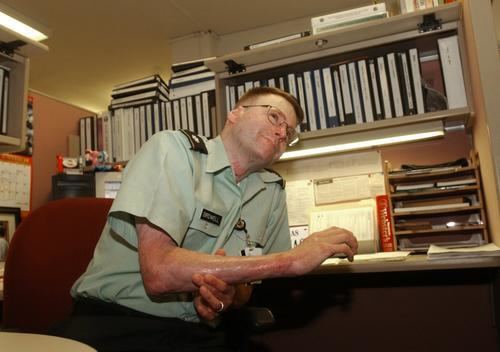 Brian Birdwell smiling while his arm was on the table and sitting on a red chair, wearing eyeglasses, a light green polo shirt uniform, and black pants.