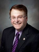 Brian Birdwell smiling, wearing eyeglasses, a black coat over purple long sleeves, and a purple tie.