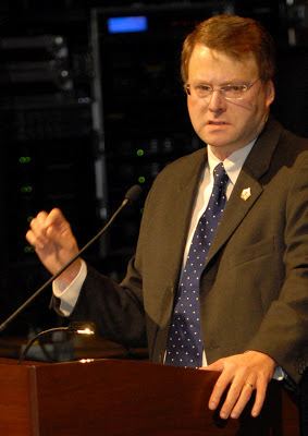 Brian Birdwell with a serious face while standing in a podium with a microphone, wearing eyeglasses, a brown coat over white long sleeves, and a blue tie.
