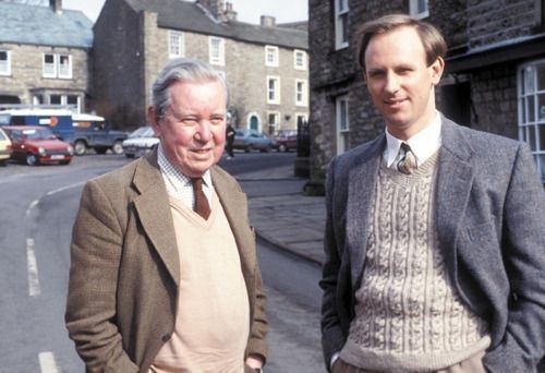 Brian Sinclair and Peter Davison are smiling while their hands are in their pockets. Brian with white hair, wearing a brown coat, a light brown vest over checkered long sleeves, and a brown tie while Peter wearing a gray coat, a light brown knitted vest over white long sleeves, and a necktie.